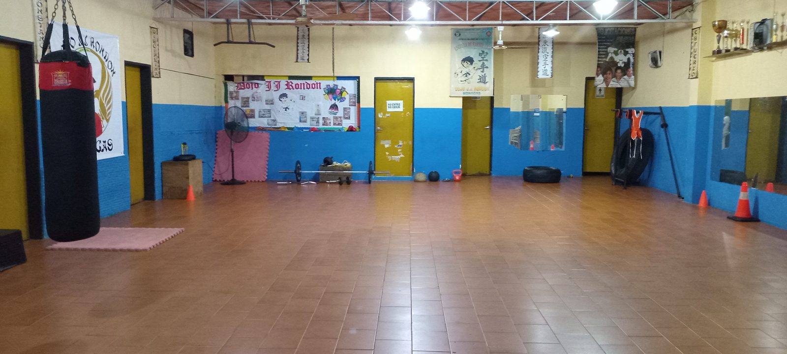 El salón principal del Dojo J.J. Rondón visto de extremo a extremo: piso de madera despejado, el saco de boxeo colgado, el mural de la escuela, la repisa de los trofeos y las llantas de entrenamiento recogidas contra la pared